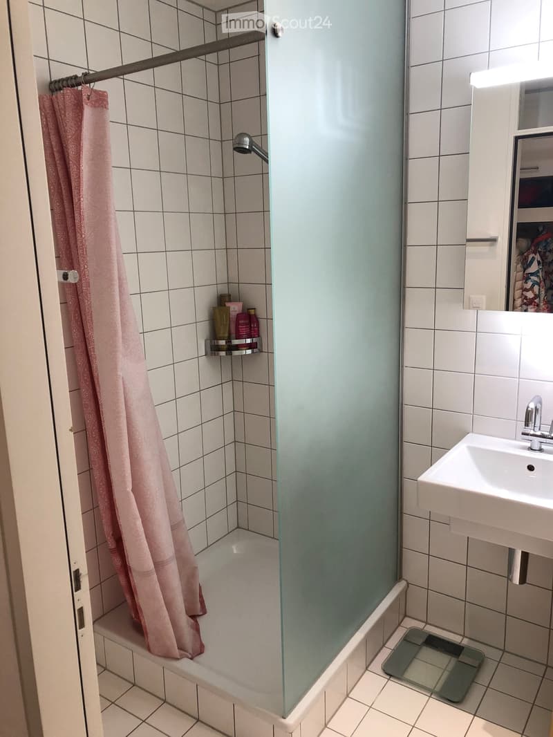 Bathroom with a bathtub, shower curtain, sink, mirror, and a small shelf holding toiletries. A glass shower enclosure is visible, and a bathroom scale is placed near the sink.
