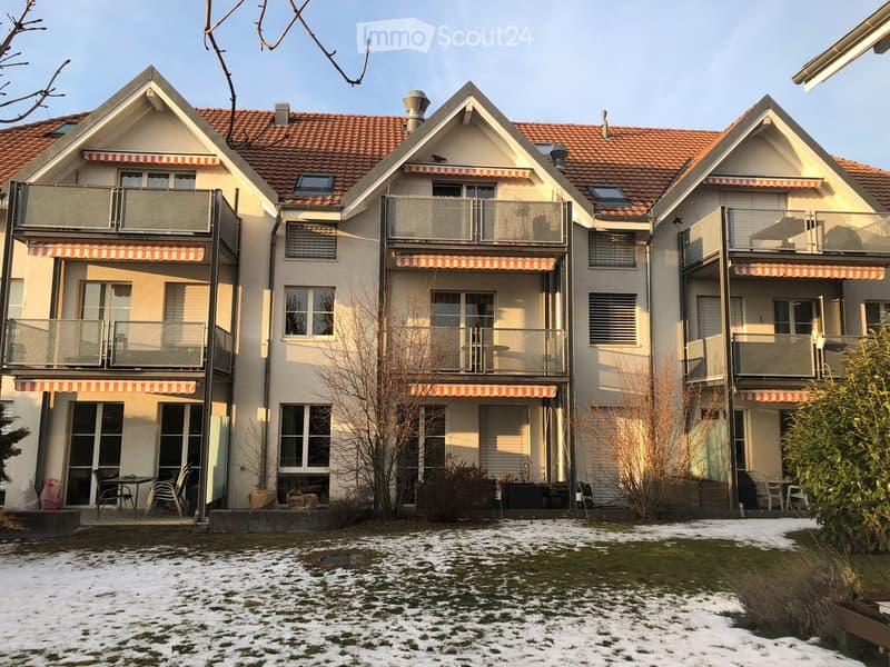 Three-story residential building with red-tiled roofs, multiple balconies, and striped awnings. The ground is partially covered in snow, and there are outdoor seating areas with tables and chairs.