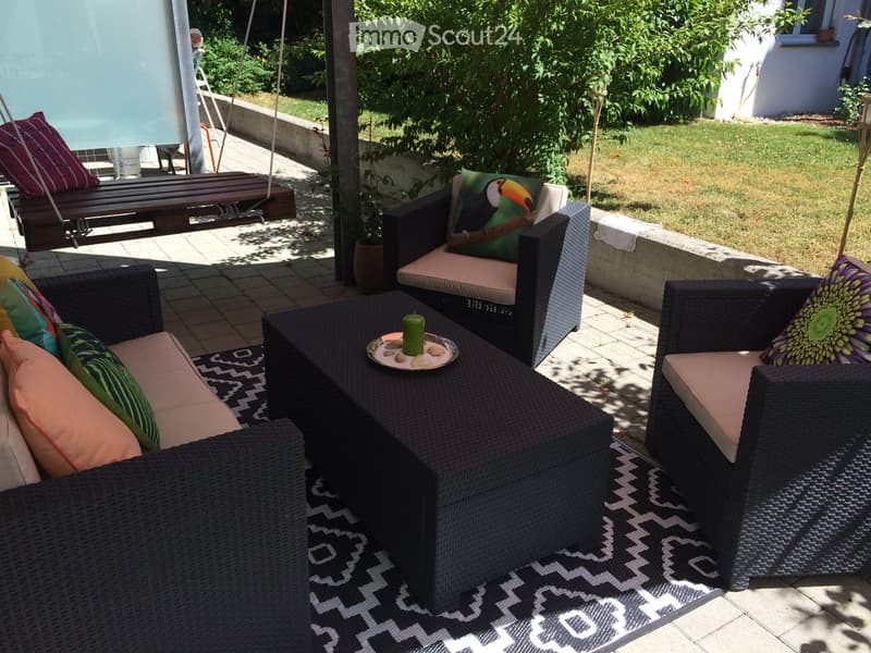 A well-furnished outdoor patio area with a sofa, chairs, a coffee table, a swing, and decorative pillows, surrounded by greenery and a swimming pool.