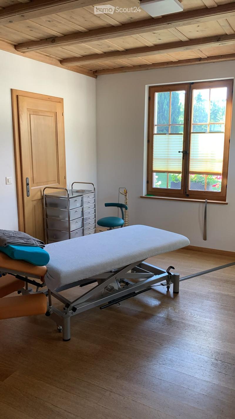 A well-lit room with wooden flooring and exposed wooden beams on the ceiling. Features a treatment table, a cabinet, a chair, and a door.