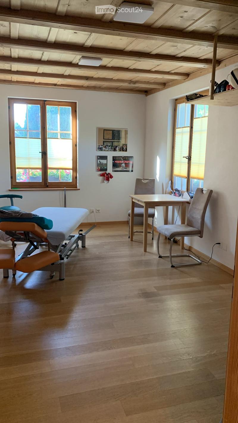 A well-lit room with wooden flooring, a treatment table, a small table with two chairs, and large windows with blinds.