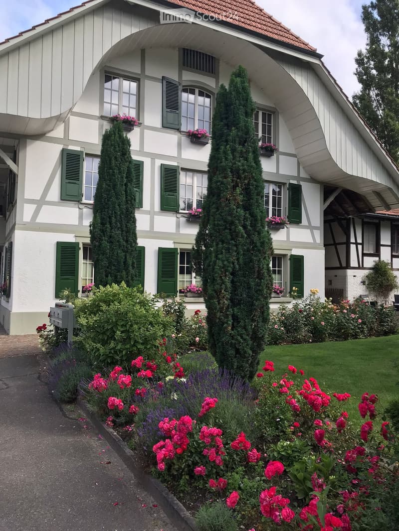 Two-story house with white and grey exterior, green shutters, and a well-maintained garden with red and pink flowers, shrubs, and tall coniferous trees.