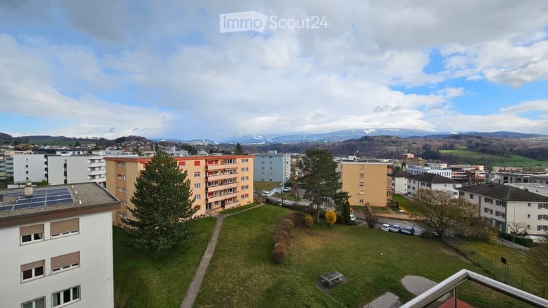 Aerial view of a residential area with multiple buildings, green grass, and trees. Solar panels on the roof of one building. Mountains in the distance.