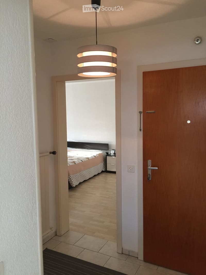 Hallway with tiled floor, wooden door, and a view into a furnished bedroom with a bed, nightstand, and lamp.