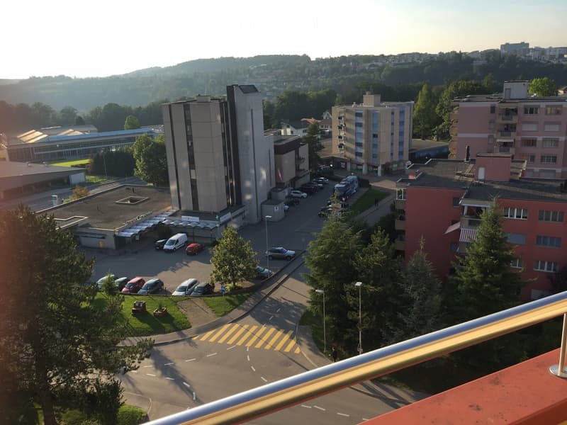 A balcony overlooking a parking lot and surrounding buildings, with a view of the city and hills in the background.