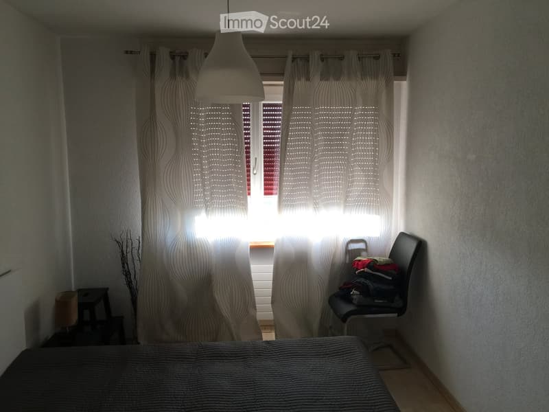 A modern bedroom with a bed, a chair with folded clothes, a small stool, and a lamp near the window with patterned curtains.