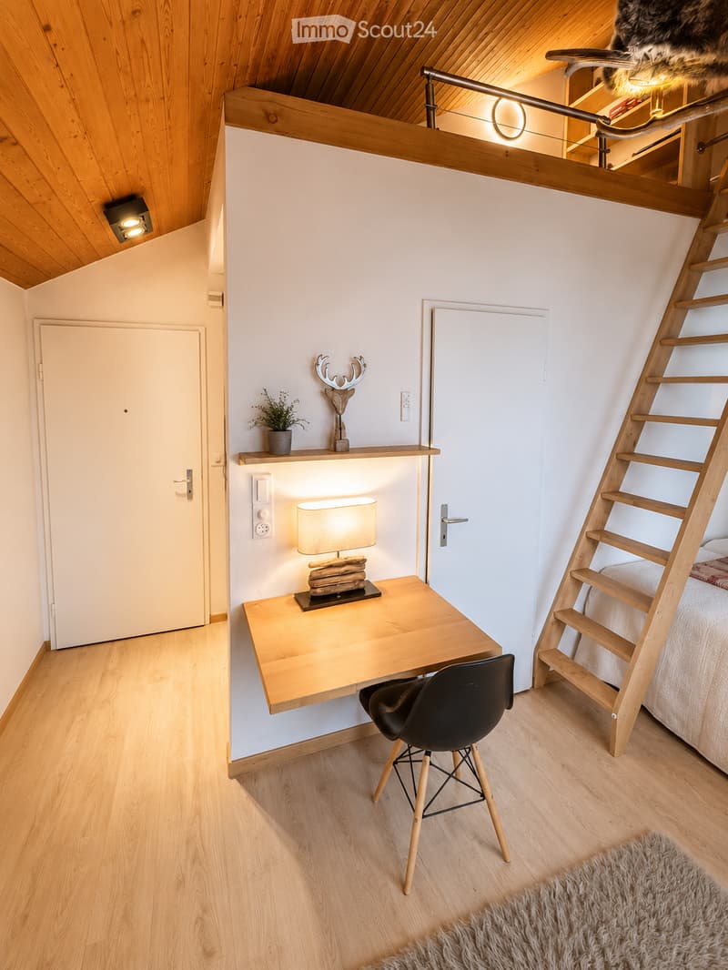 Cozy attic bedroom with wooden floors, slanted wooden ceiling, built-in desk, chair, lamp, small shelf with plant and deer head, and a wooden ladder leading to a lofted sleeping area.