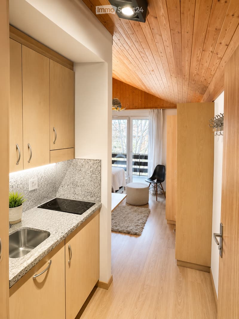 Modern kitchen with light wood cabinets, granite countertop, induction stove, and sink. Adjacent living area features a round stool, chair, and a small rug.
