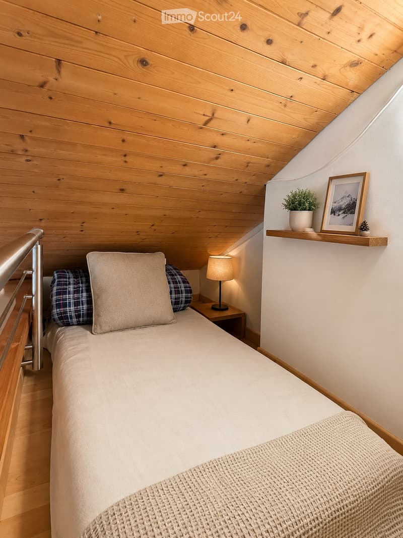 Cozy bedroom with a single bed, wooden ceiling, nightstand with a lamp, and a small shelf with a picture frame and potted plant.