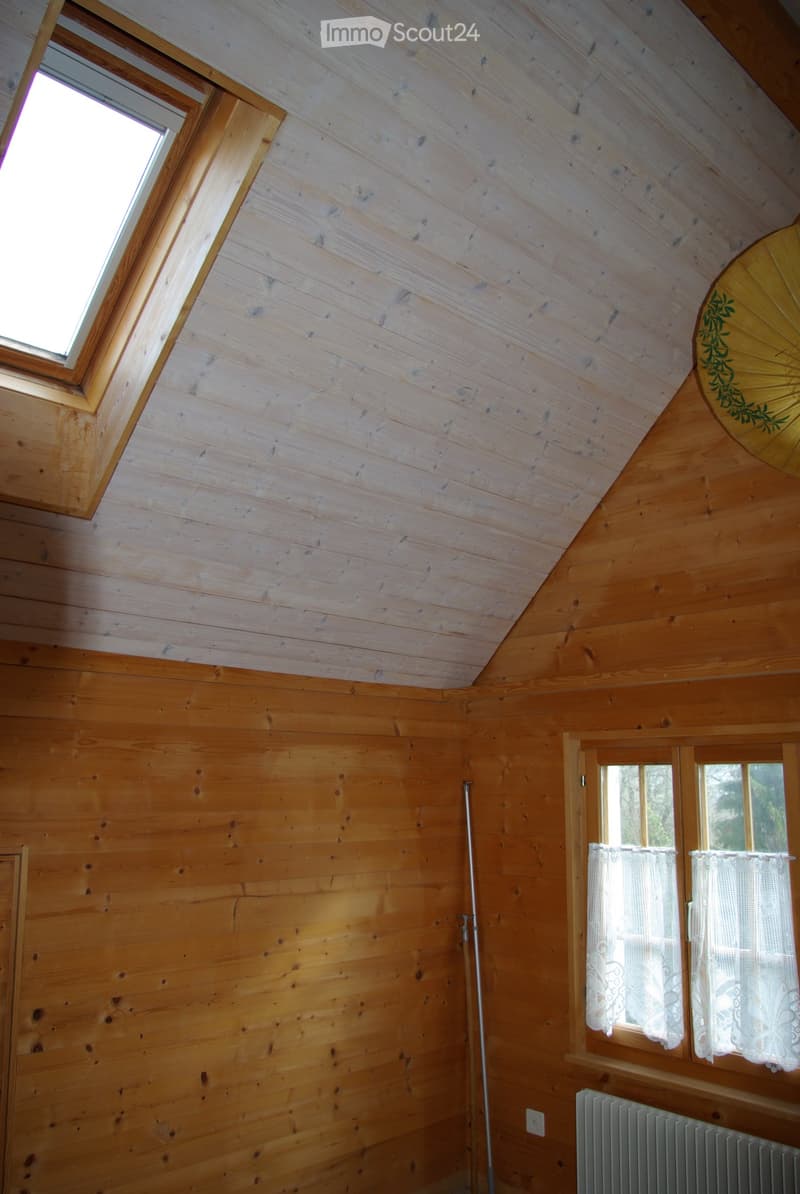 Empty wooden room with slanted ceiling, skylight window, and two side windows with white curtains. Wooden walls and floor, radiator, and a floor lamp stand visible.