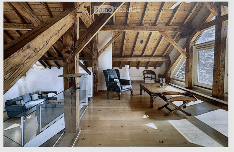 Loft-style living room with exposed wooden beams and high ceilings. Features a grey sofa, armchair, wooden table, and a stool. Large windows allow natural light to fill the space.