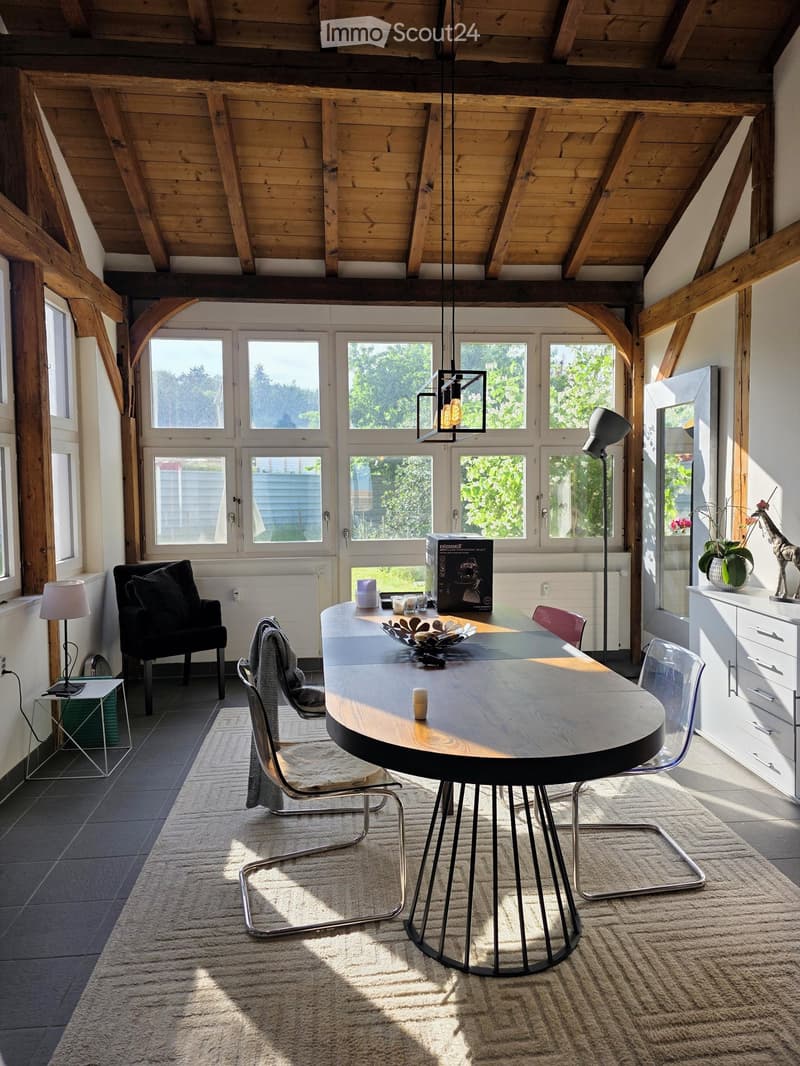 Bright, modern living space with exposed wooden beams, large windows, a round wooden table with chairs, a black sofa, a side table with a lamp, and a decorative cabinet with a vase and figurine.