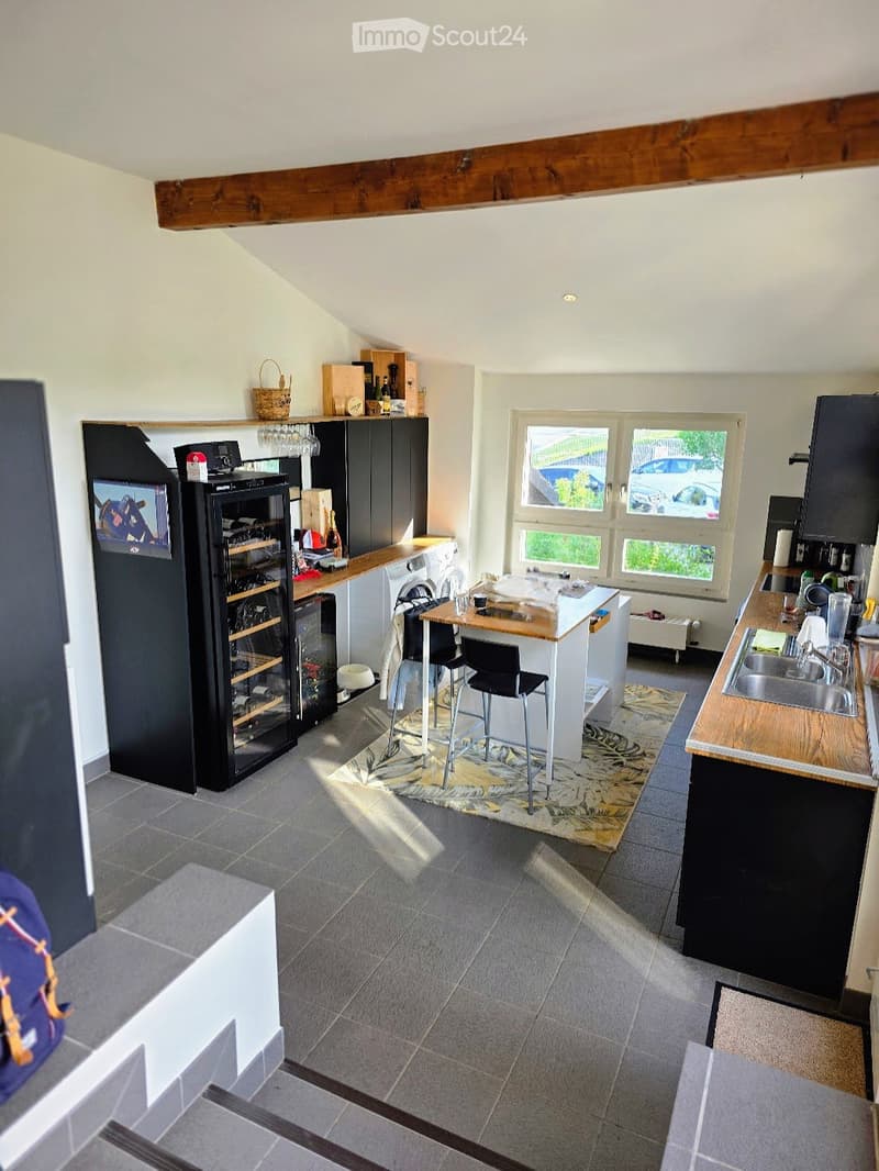 Modern kitchen with grey tiled floor, white walls, and dark cabinets. Features a wine cooler, a small table with stools, a sink with a faucet, and various kitchen items on the counters.