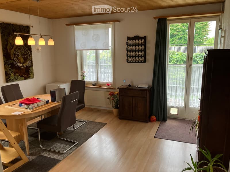 Brightly lit room with wooden flooring, a round wooden table surrounded by chairs, a dark wooden cabinet, and a large window with blinds. There is a decorative wall piece and a small shelf with jars visible.