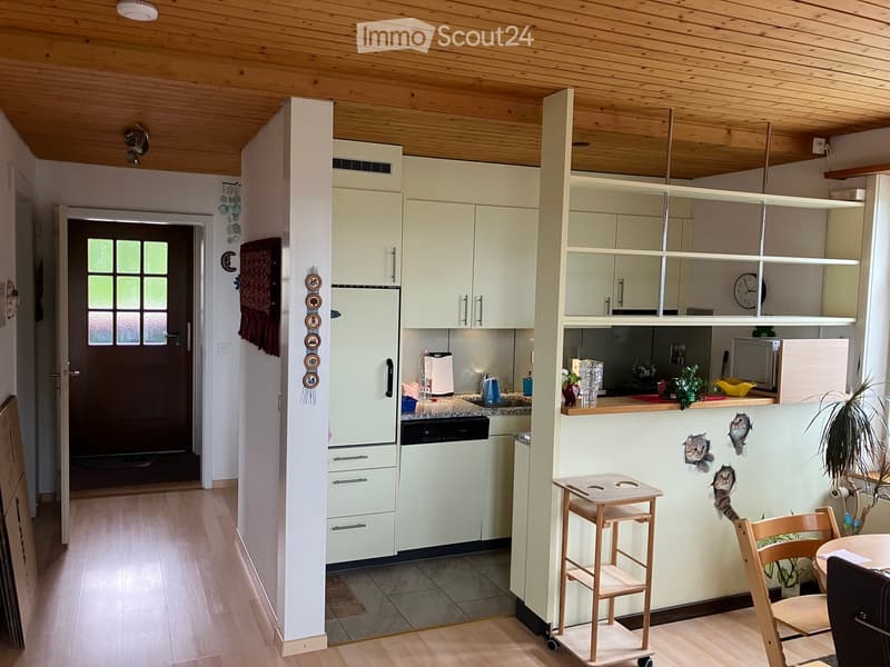 Bright kitchen with light-colored cabinets, a refrigerator, dishwasher, microwave, and a small wooden stool. There is a small table with a chair, a potted plant, and a cat sticker on the wall.