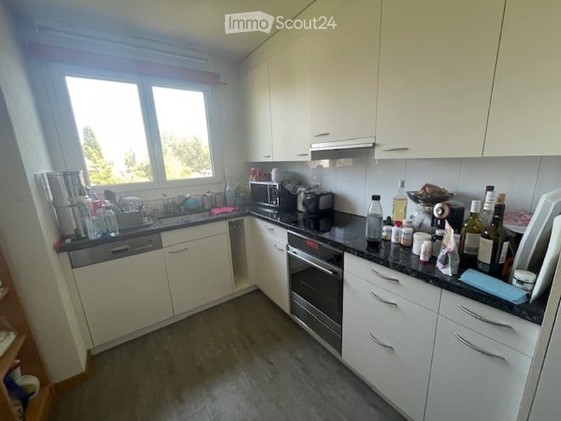 Modern kitchen with white cabinets, black countertops, and various appliances including a dishwasher, oven, microwave, and coffee maker. The countertop is cluttered with bottles, cups, and other kitchen items.