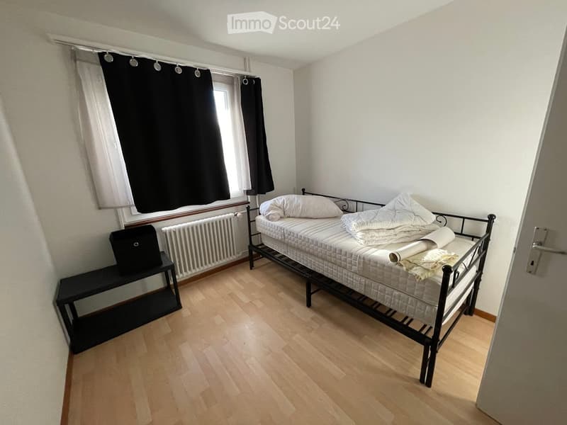 Bedroom with a black frame bed, white bed sheets, pillows, a nightstand, white radiator, black curtains, and wooden floors.