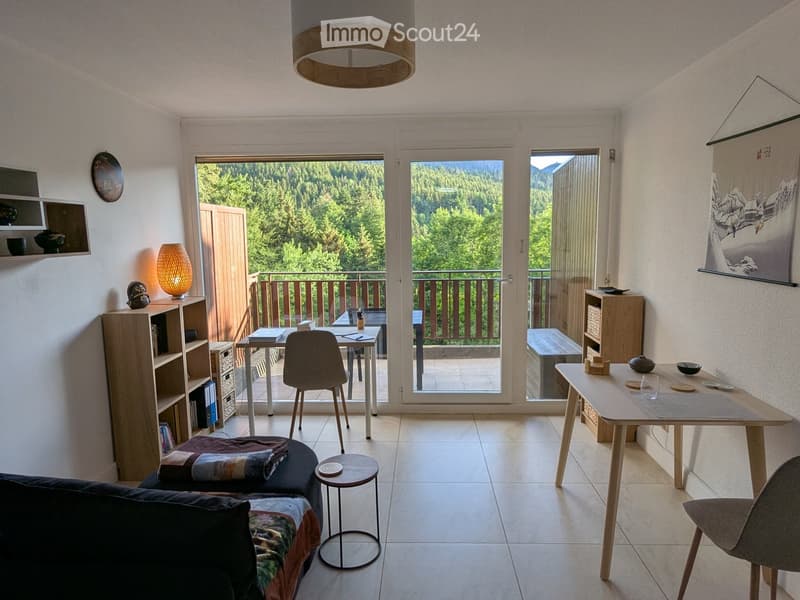 Modern living room with a sofa, small round table, dining table, chairs, shelves, and a view of the forest through large windows and a balcony.