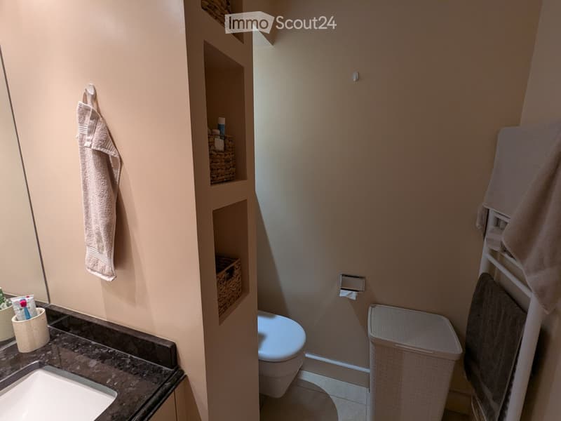 A small, modern bathroom with a sink, toilet, and built-in wicker baskets for storage. Towels are neatly hung on racks, and a trash bin is placed near the toilet.