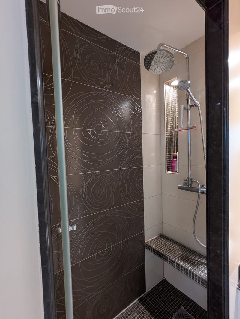 Modern shower area with dark brown tiles featuring abstract swirls, a glass door, a built-in shelf holding a bottle, and a multi-function showerhead with handheld sprayer.