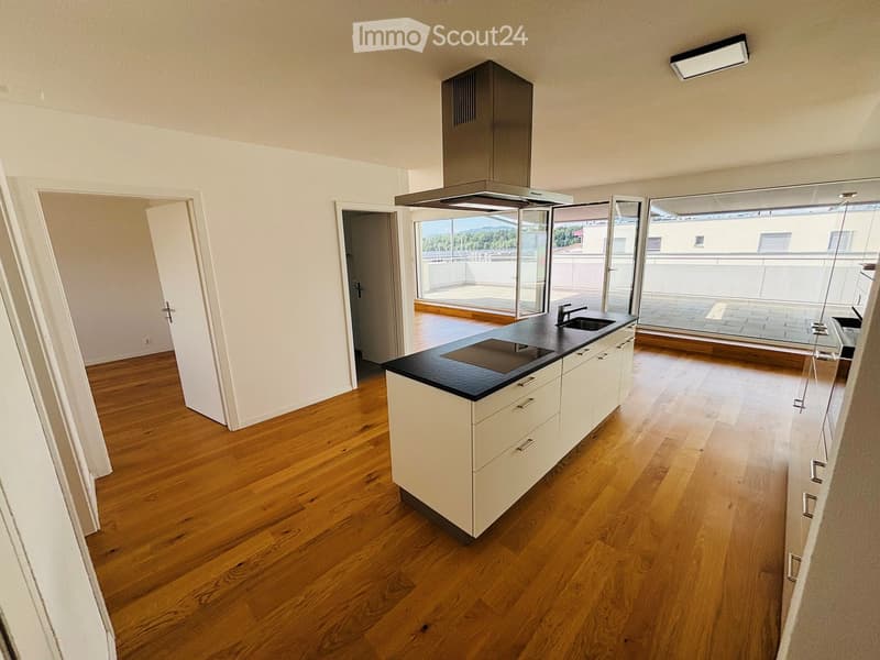 Modern kitchen with light-colored cabinets, a black countertop, and a range hood. The space features wooden flooring and large windows offering a scenic view.
