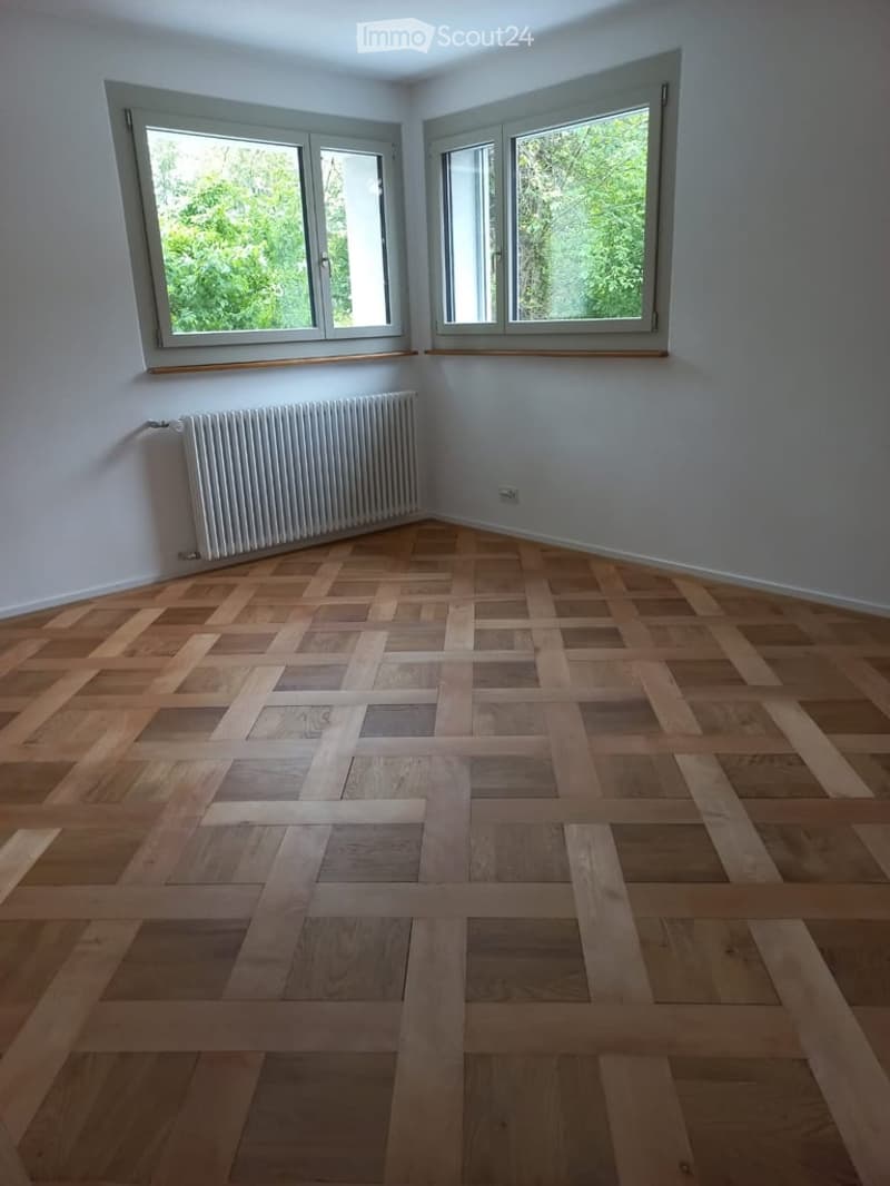 Empty room with wooden herringbone flooring, white walls, and two large windows with white frames. A white radiator is mounted on one wall.