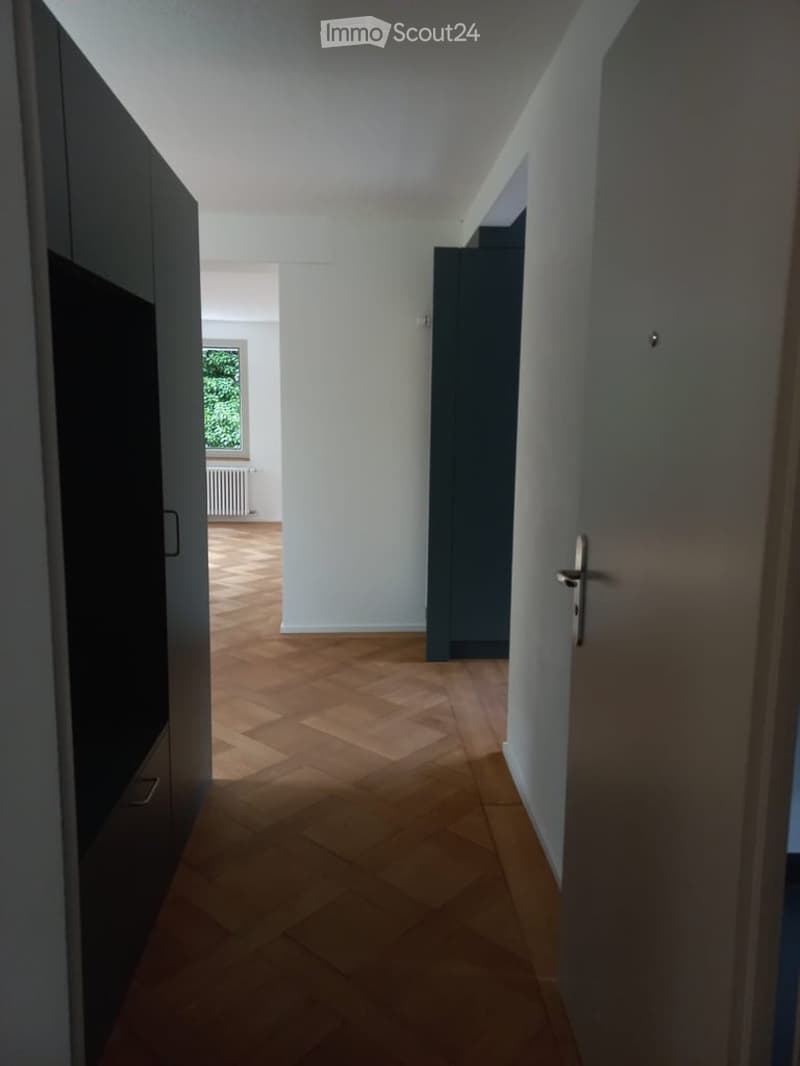 A narrow hallway with wooden herringbone flooring, white walls, and dark grey doors on both sides. Natural light filters in from a window at the end of the corridor.