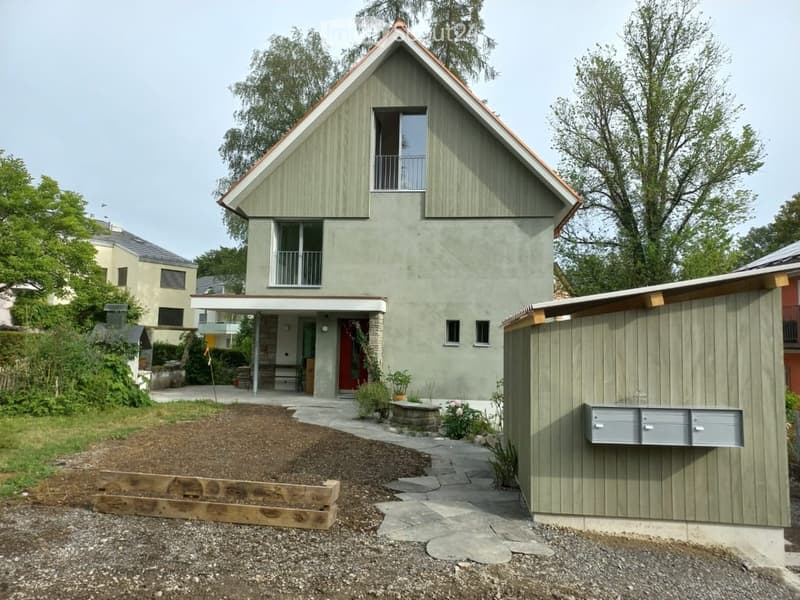 Modern two-story house with green siding, red door, and a small porch. The house features a raised ground floor, a small garden area, and a wooden planter box in the front yard.