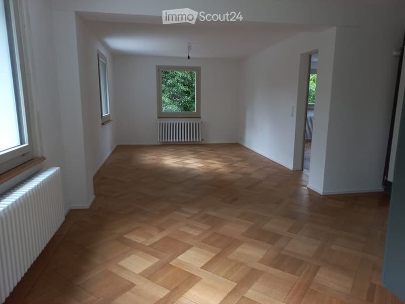 Empty room with wooden herringbone flooring, white walls, and large windows allowing natural light. Radiators are visible along the walls.