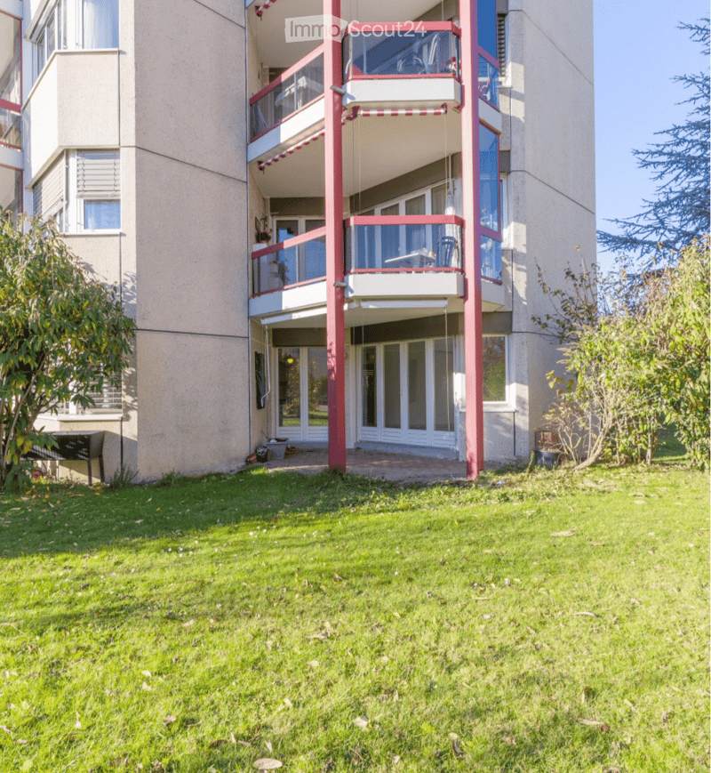 Multi-story residential building with balconies, glass railings, and a well-maintained lawn in front.