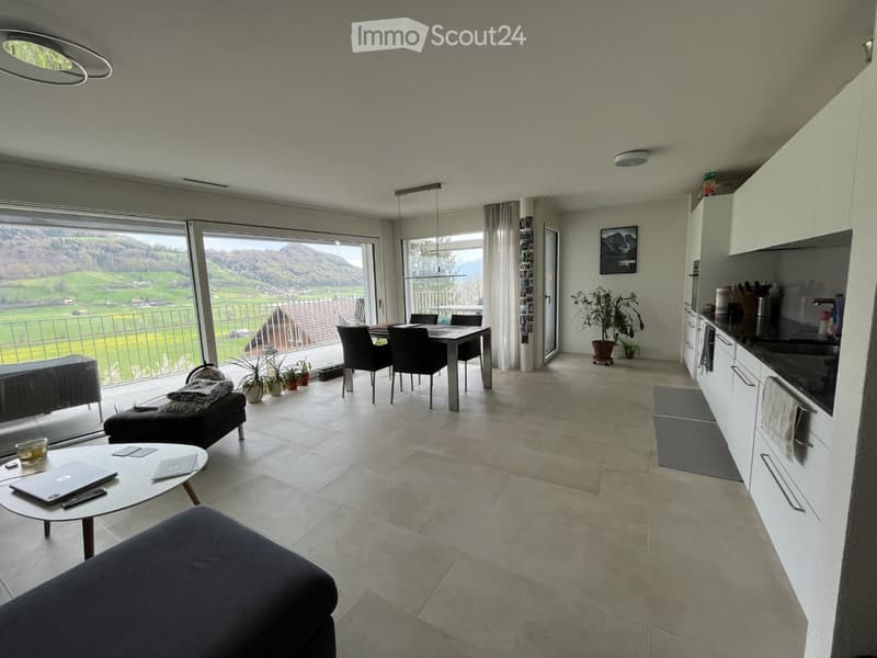 Open-plan living and dining area with large windows offering a scenic view, featuring a sofa, coffee table, dining table, chairs, and a modern kitchen area.