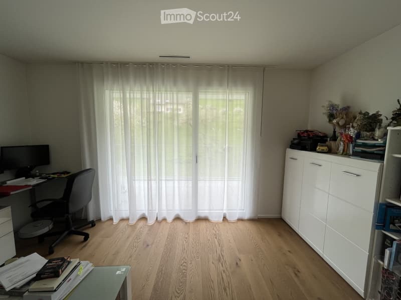 A well-lit room with wooden flooring, a large window with white curtains, a desk with a computer and chair, a white cabinet, and a shelf with books and decorative items.