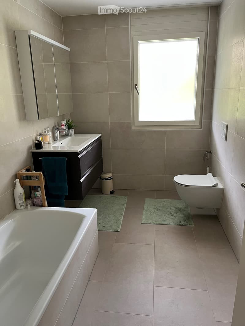 Modern bathroom with beige tiles, a bathtub, toilet, sink with mirror, and a small plant on the sink counter.