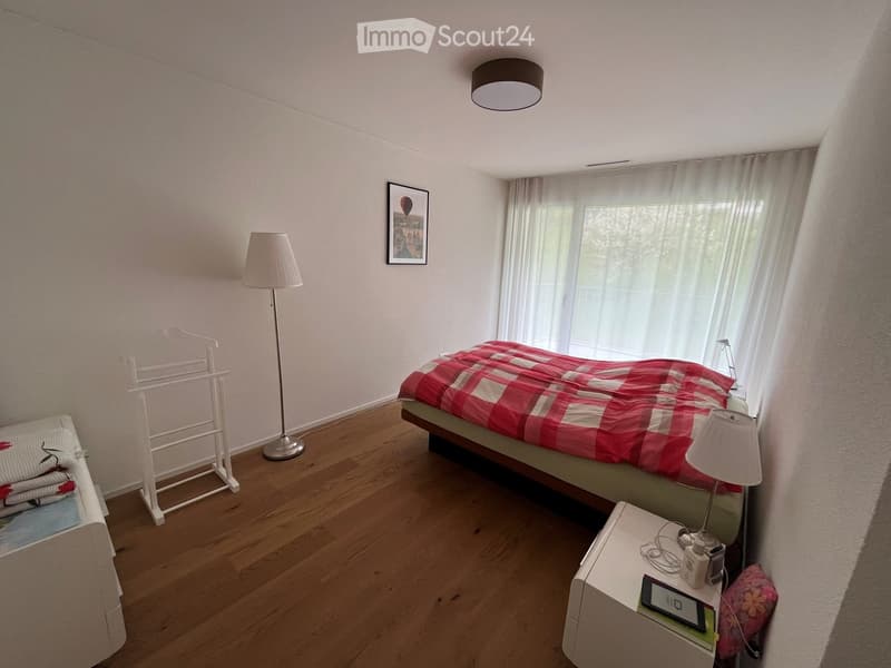Cozy bedroom with wooden flooring, a single bed with red and white checkered bedding, a nightstand with a lamp, a picture frame on the wall, and a clothing rack with folded towels.