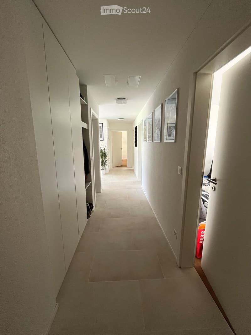 Clean, well-lit hallway with beige tiled flooring, white walls, and recessed ceiling lighting. Features built-in shelving on one side and framed artwork on the other, with a small plant visible in the distance.
