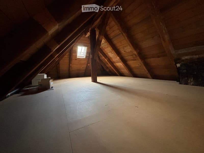 Empty attic space with wooden beams, exposed rafters, and a small window letting in natural light. The floor is unfinished concrete.