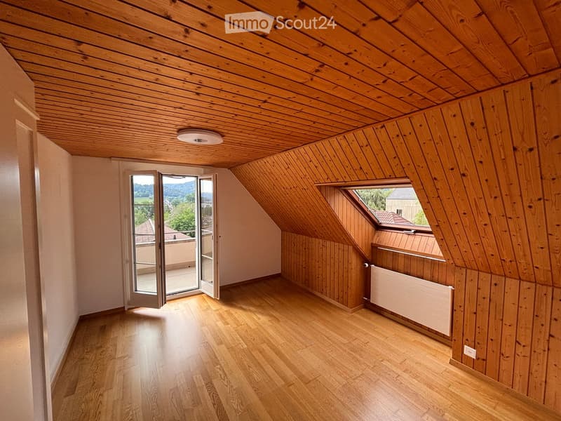 Bright, empty attic room with wooden walls, ceiling, and floor, featuring a large window and a sliding glass door leading to a balcony.