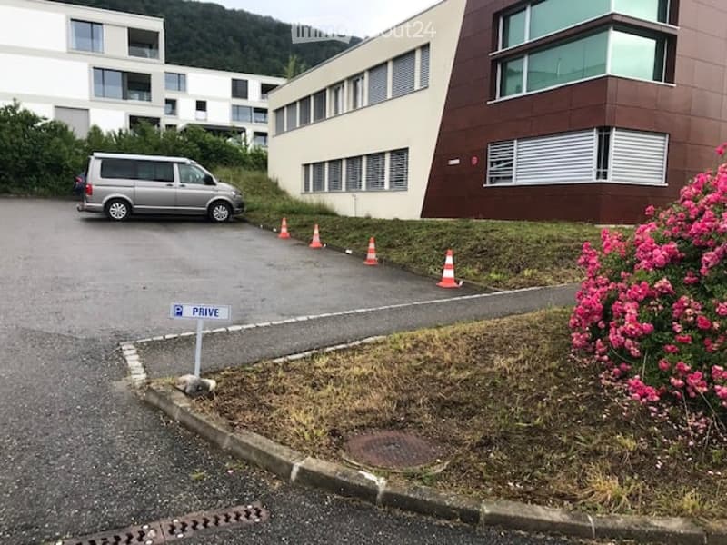 Parking area with a silver van, traffic cones, and a sign reading "PRIVE" near a modern building with large windows and a landscaped garden with pink flowers.