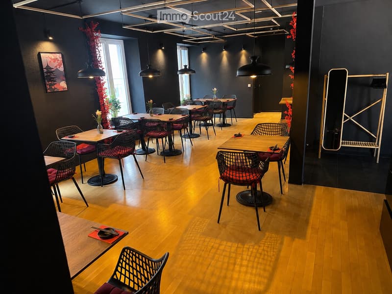 Modern restaurant interior with wooden floors, black walls, and pendant lighting. Tables and chairs are arranged throughout, with decorative vases and artwork on the walls.
