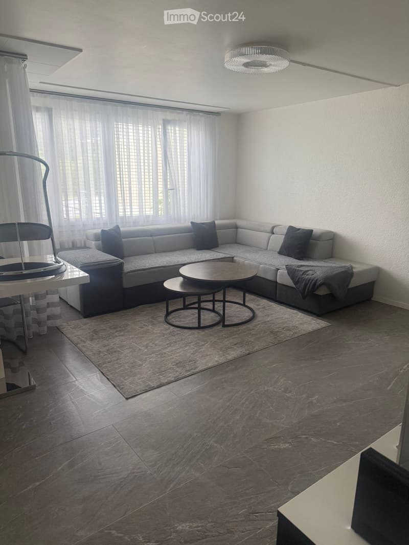 Modern living room with a grey sectional sofa, coffee table, side table, and a large window with sheer curtains. The room features a marble floor and a contemporary chandelier.