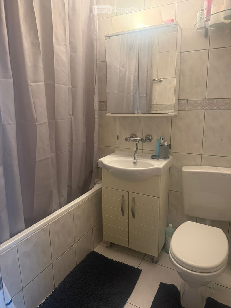 A small bathroom with a white sink, faucet, toilet, and a mirrored cabinet above the sink. The floor has beige tiles, and there are black rugs placed in front of the sink and toilet.