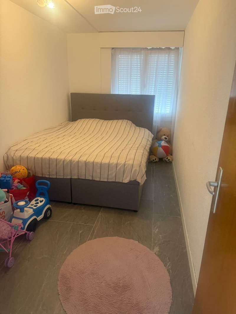 A cozy bedroom with a bed, a round pink rug, and various toys including a toy car, a stroller, a teddy bear, and a ball. The room has a window with blinds and a door visible.