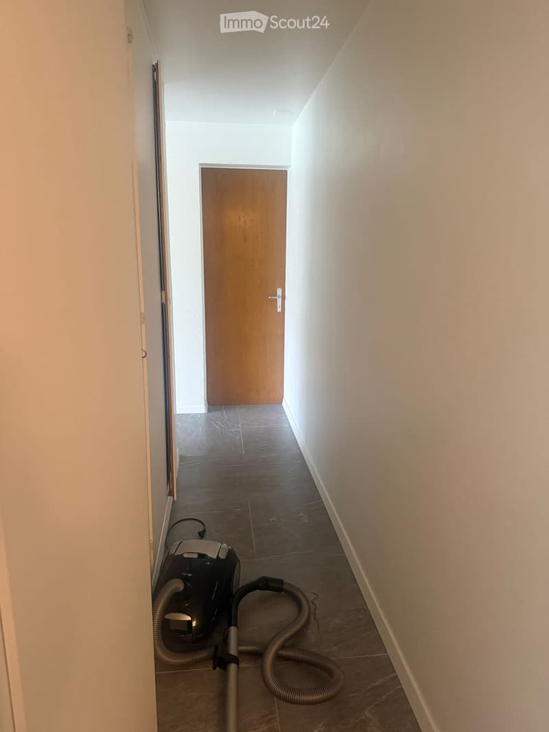 Empty hallway with light-colored walls, tiled floor, and a closed wooden door at the end. A vacuum cleaner is placed on the floor near the left wall.