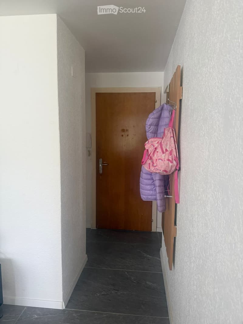 A narrow hallway with a dark tiled floor, white walls, and a wooden door at the end. A coat rack with a purple jacket and a pink backpack is visible on the right side.