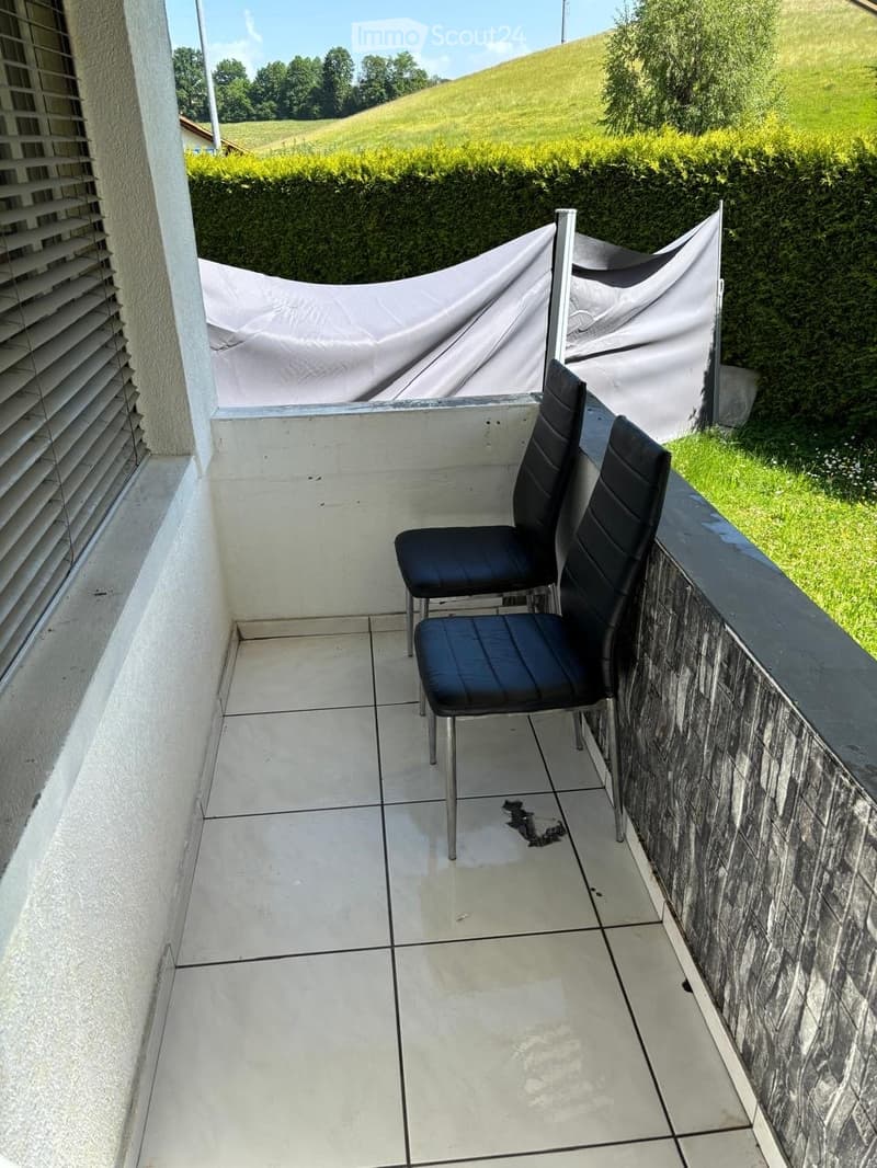 Balcony with tiled floor, two black chairs, covered area with white fabric, greenery in the background.