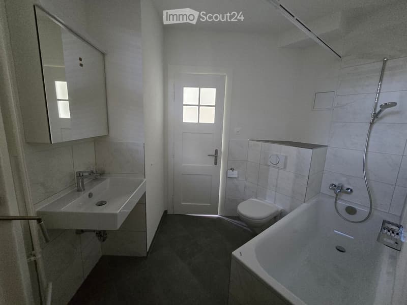 Modern bathroom with a white sink, mirror, toilet, bathtub, and showerhead. The room features a white door with a window and a small cabinet above the sink.