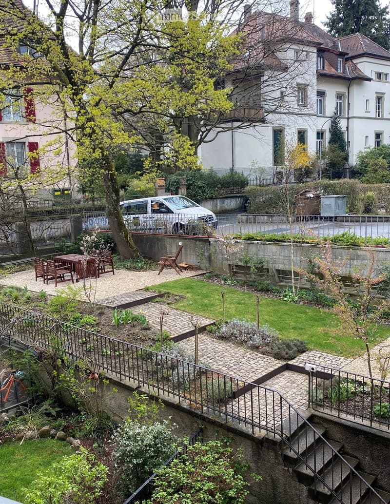 Garden with a raised ground floor, featuring a small lawn, gravel paths, a wooden table with chairs, and a bench. There is a staircase leading down to a lower garden area with more plants and a metal railing.