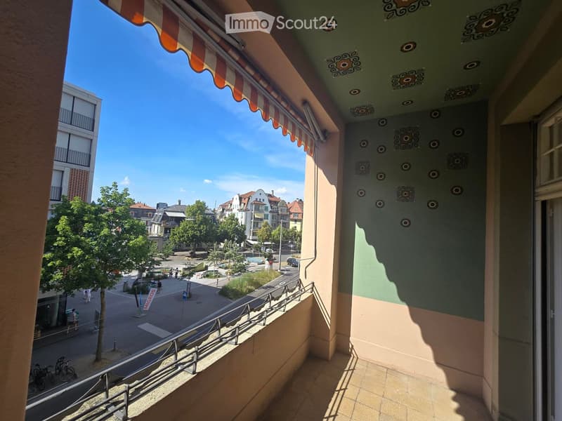 Balcony with striped awning, metal railing, and view of a street with trees, buildings, and pedestrians.