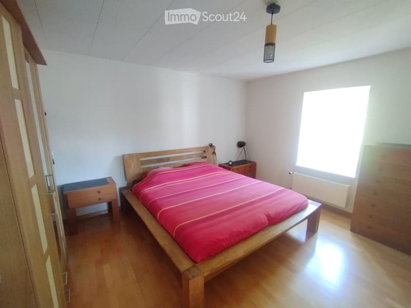 Bright bedroom with wooden flooring, a wooden bed with red bedding, a nightstand with a lamp, and a dresser.