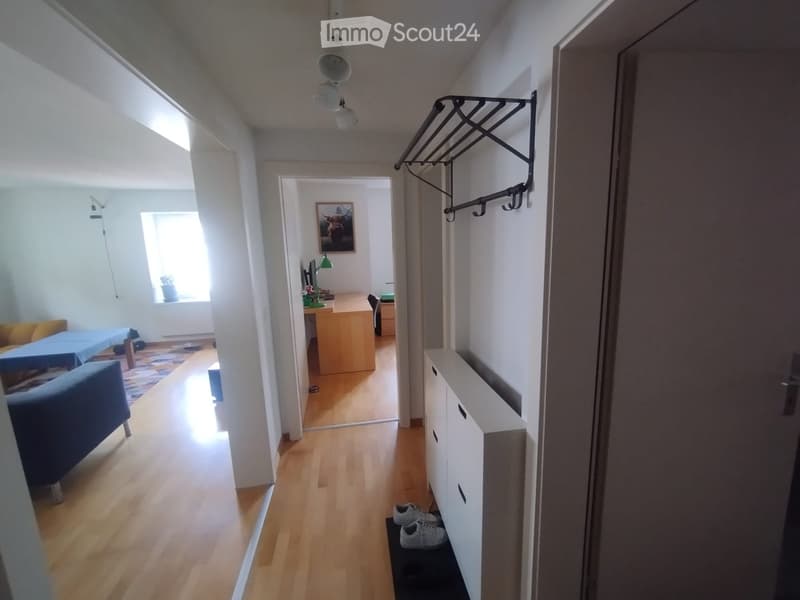 Bright hallway with wooden flooring, white walls, and a coat rack. Visible rooms include a living area with a sofa and a study with a desk and chair.